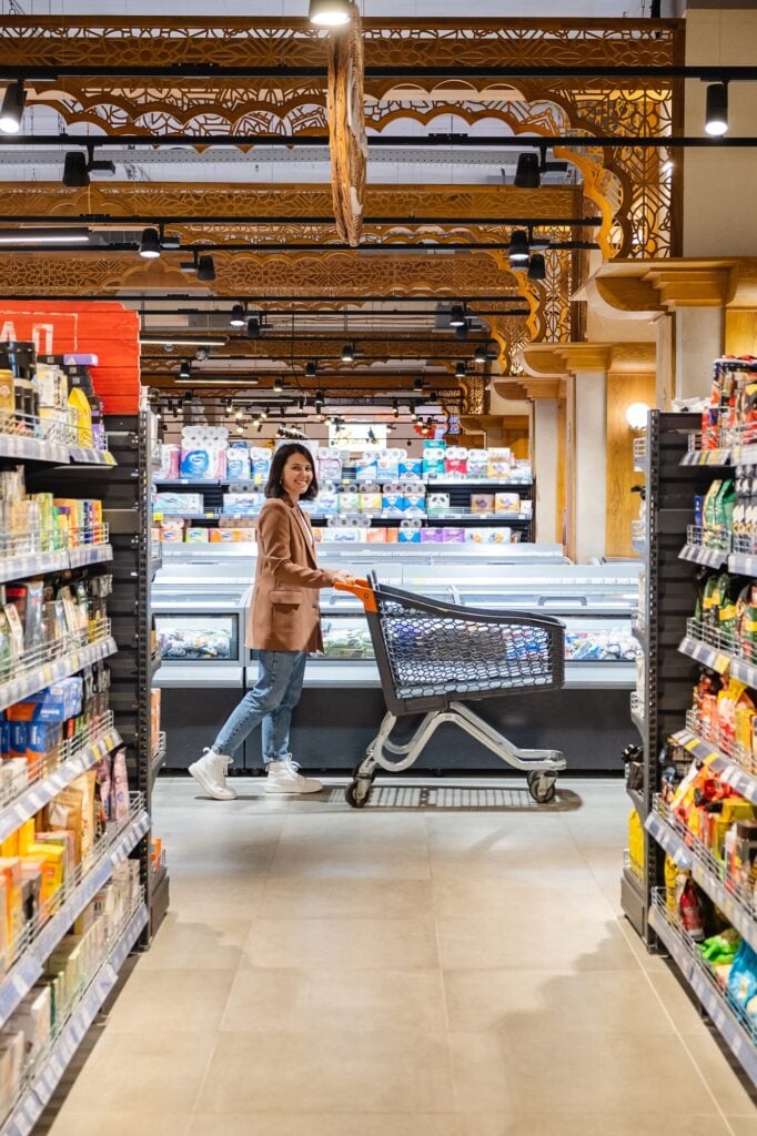 a woman with a cart walks between rows of shelves in a grocery store